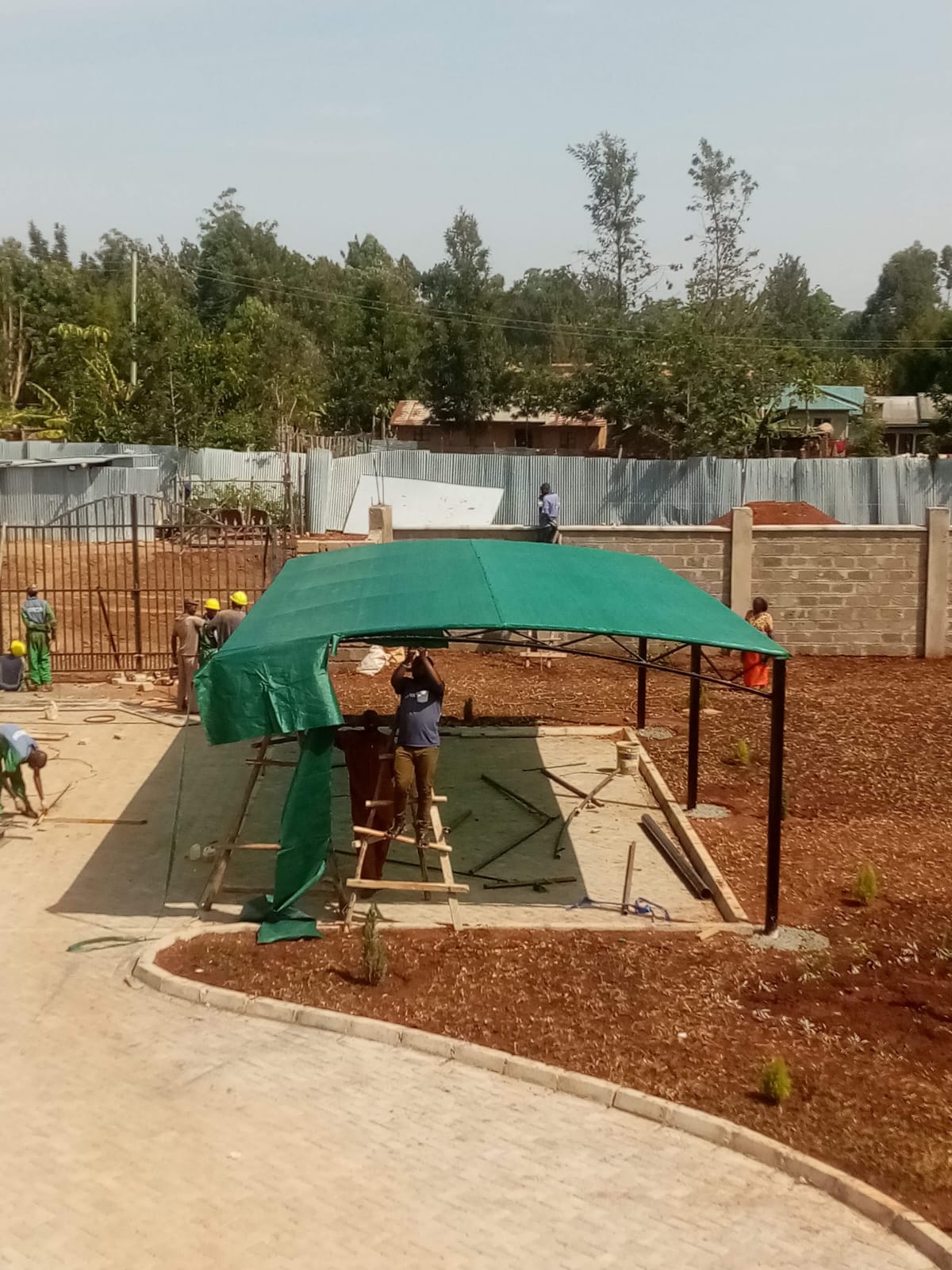 Technicians fitting green tensile fabric to a curved car parking shade frame in Bungoma, with a walled compound and eucalyptus trees behind
