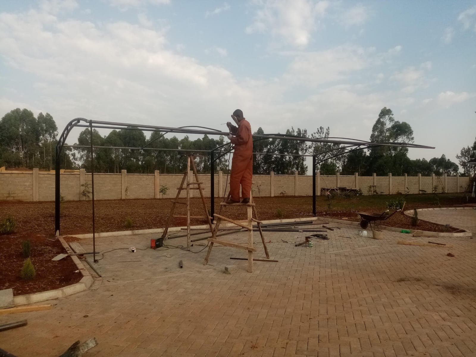 Technician welding a curved steel cantilever frame for a car parking shade structure in Bungoma, standing on a wooden ladder
