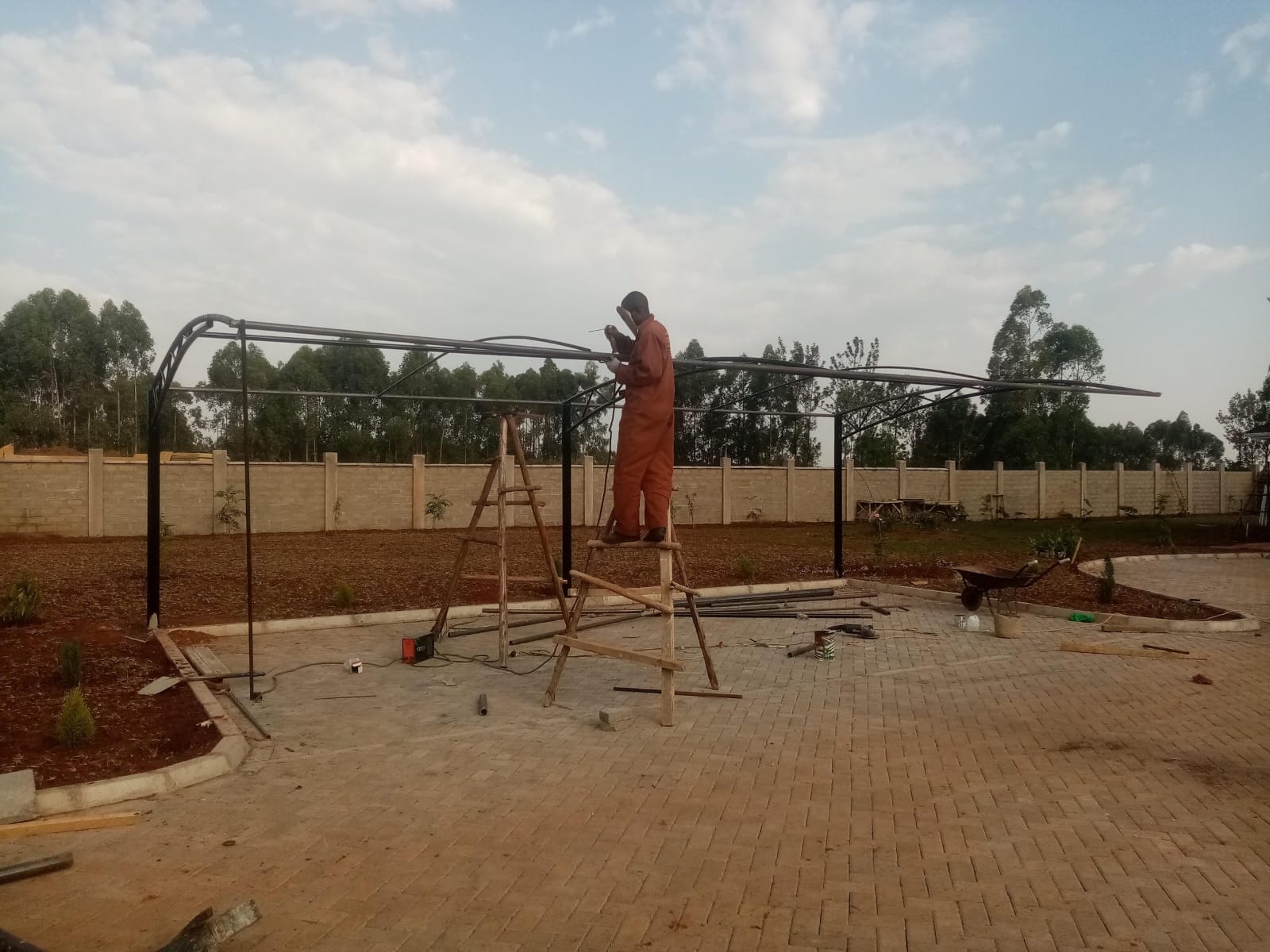 Technician welding a curved steel cantilever frame for a car parking shade structure in Bungoma, standing on a wooden ladder