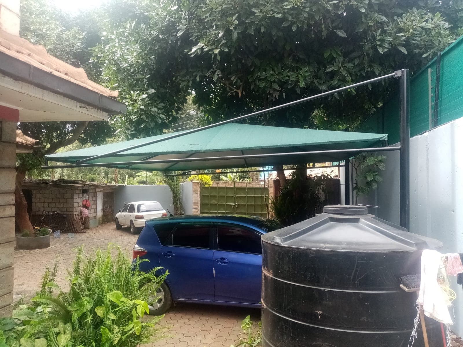 Green single-post cantilever car parking shade structure over a residential driveway in Kikuyu, beneath mature trees alongside a boundary wall