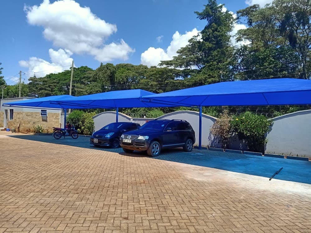 Blue tensile cantilever car parking shade structure covering vehicles along a stone-clad boundary wall in Nakuru