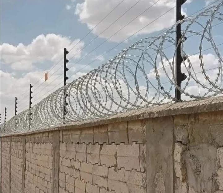 Silver razor wire coils and electric fence wire strands installed along a stone-block boundary wall in Garissa