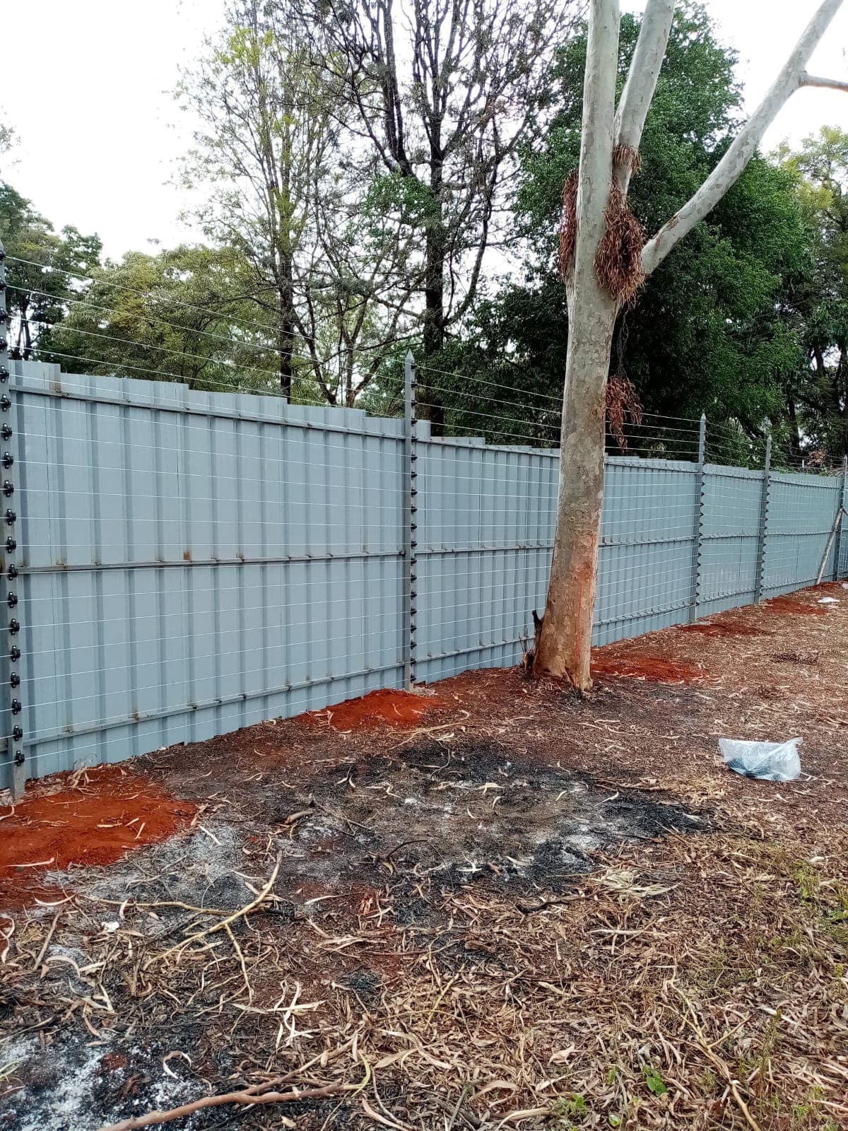 Multi-strand electric fence wires running along a light blue corrugated boundary wall in Karen, Nairobi, under mature gum trees