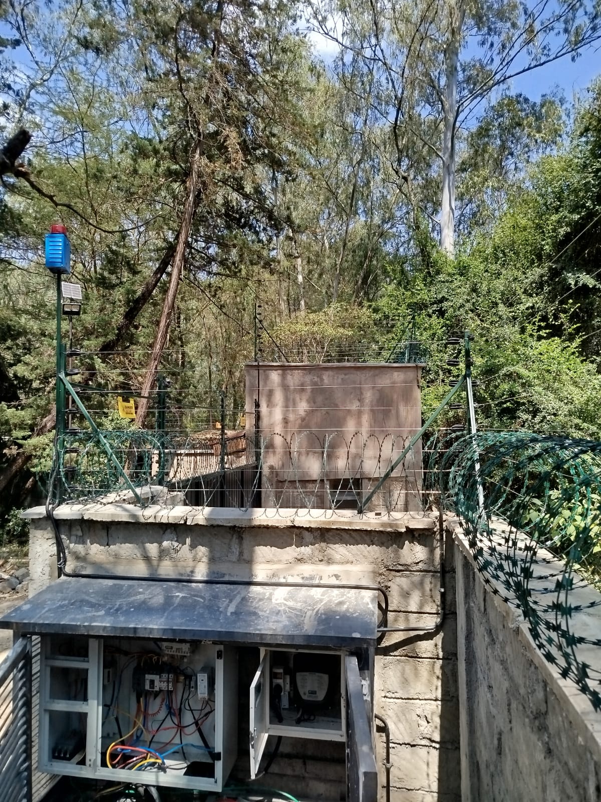 Solar-powered siren mounted on a boundary wall post above razor wire and electric fence wiring in Karen woodland