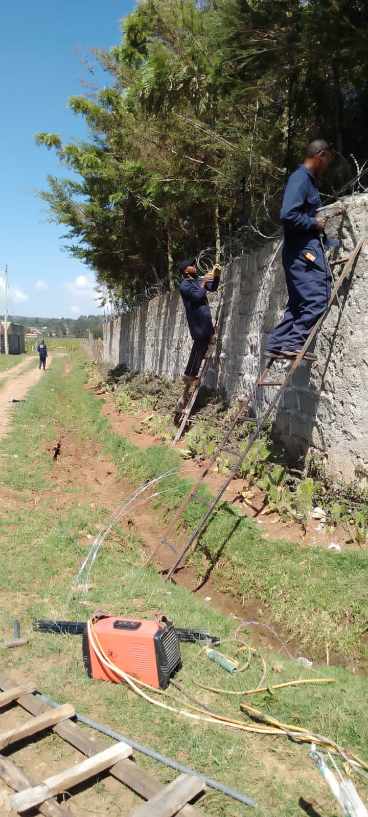 Two technicians on ladders fitting razor wire and electric fence posts along a boundary wall in Nanyuki, with a welding machine on the ground