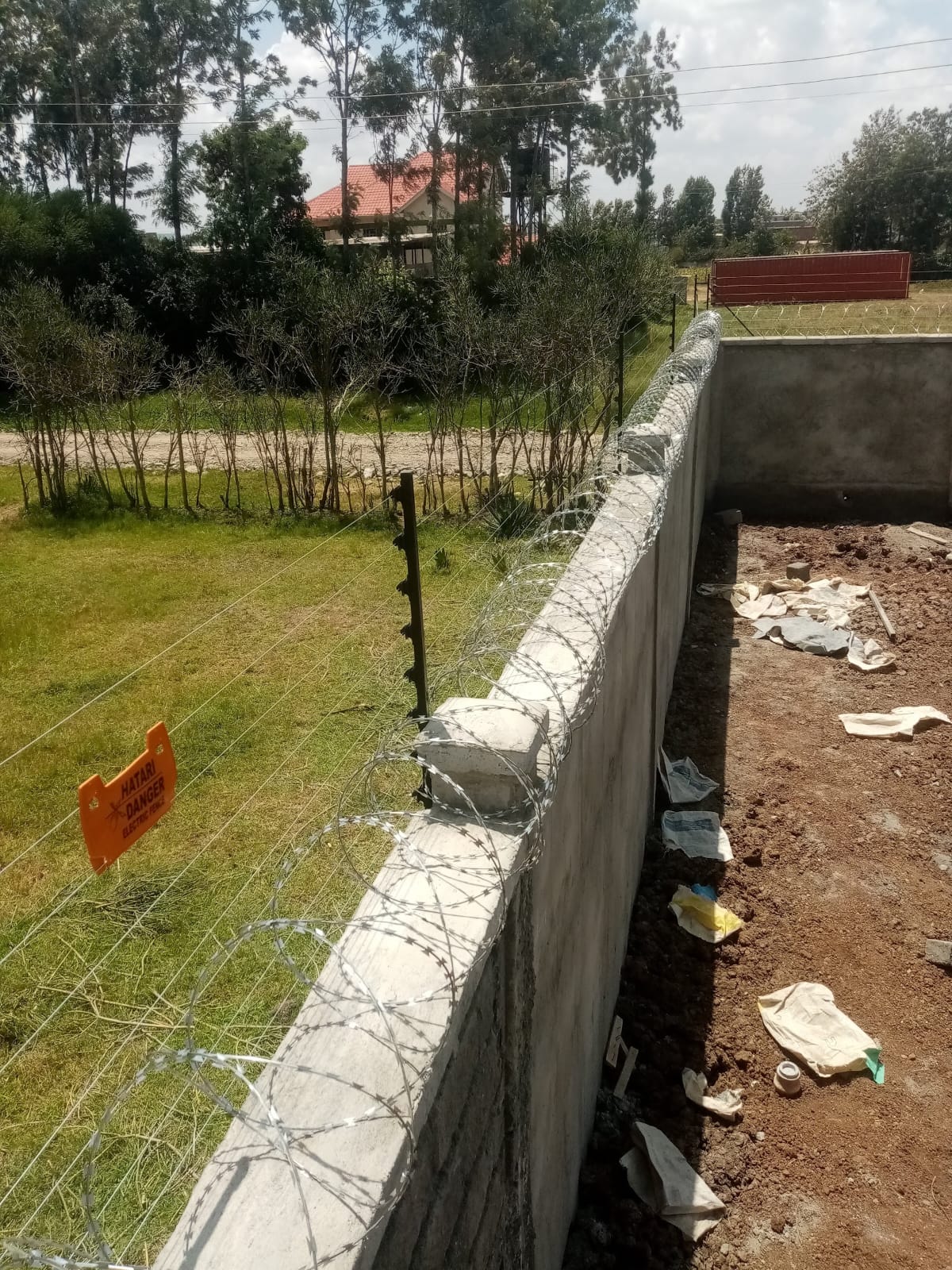 Electric fence and razor wire along a newly built boundary wall in Ruiru, with a red-roofed house visible beyond the perimeter