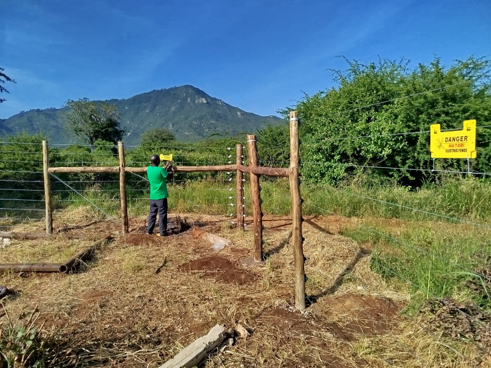 Technician mounting a Hatari electric fence danger sign on a timber post near Voi, with Tsavo hills behind