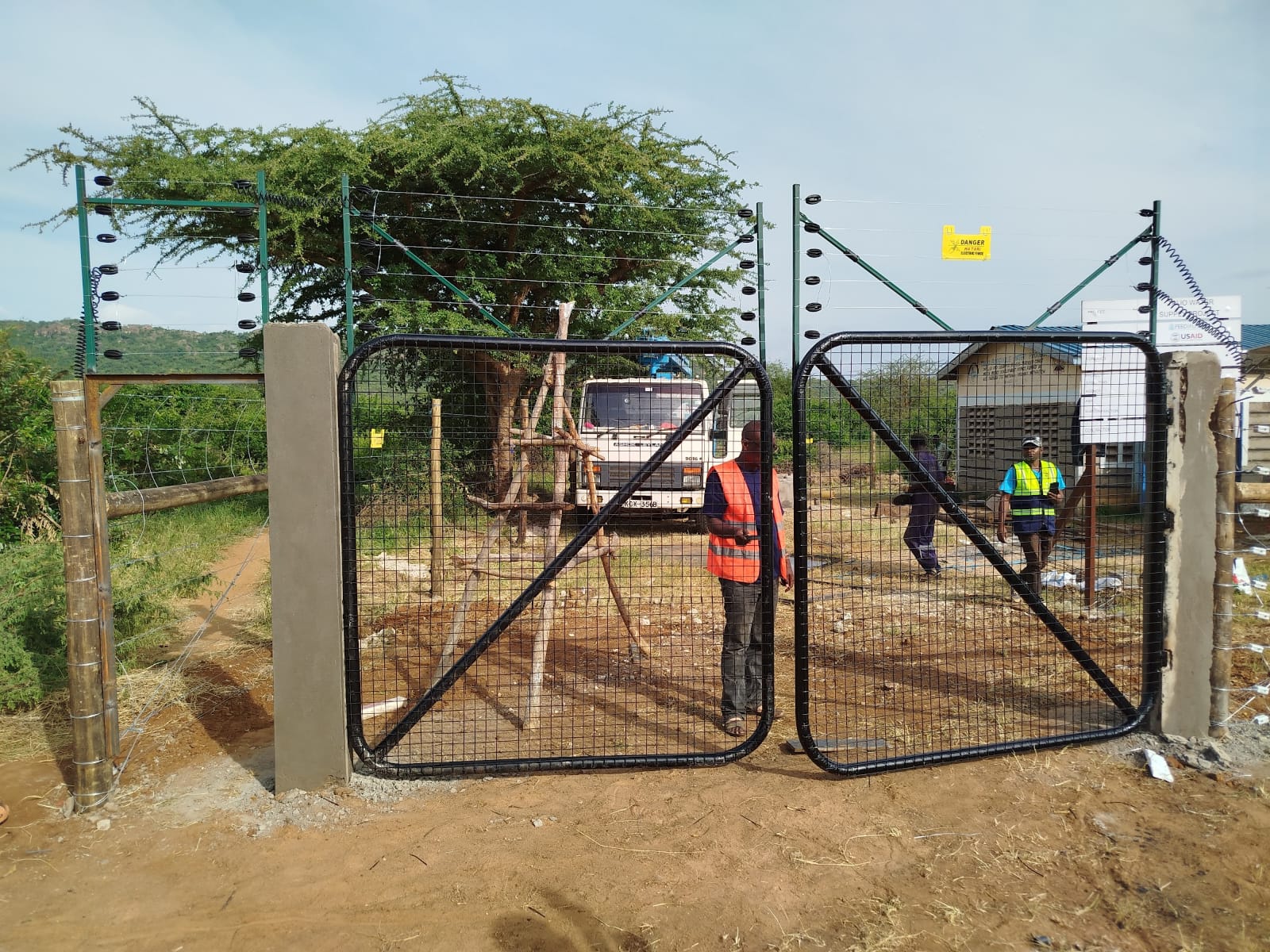 Finished double mesh gate topped with electric fence at an institutional site near Voi, hi-vis workers and USAID signage visible