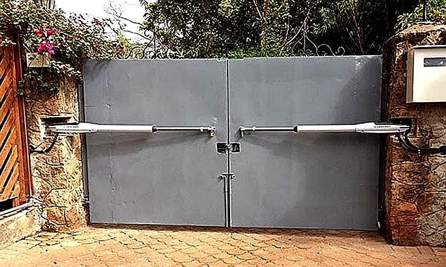 Grey double-leaf solid swing gate with dual arm motor actuators installed between stone entrance pillars in Karen