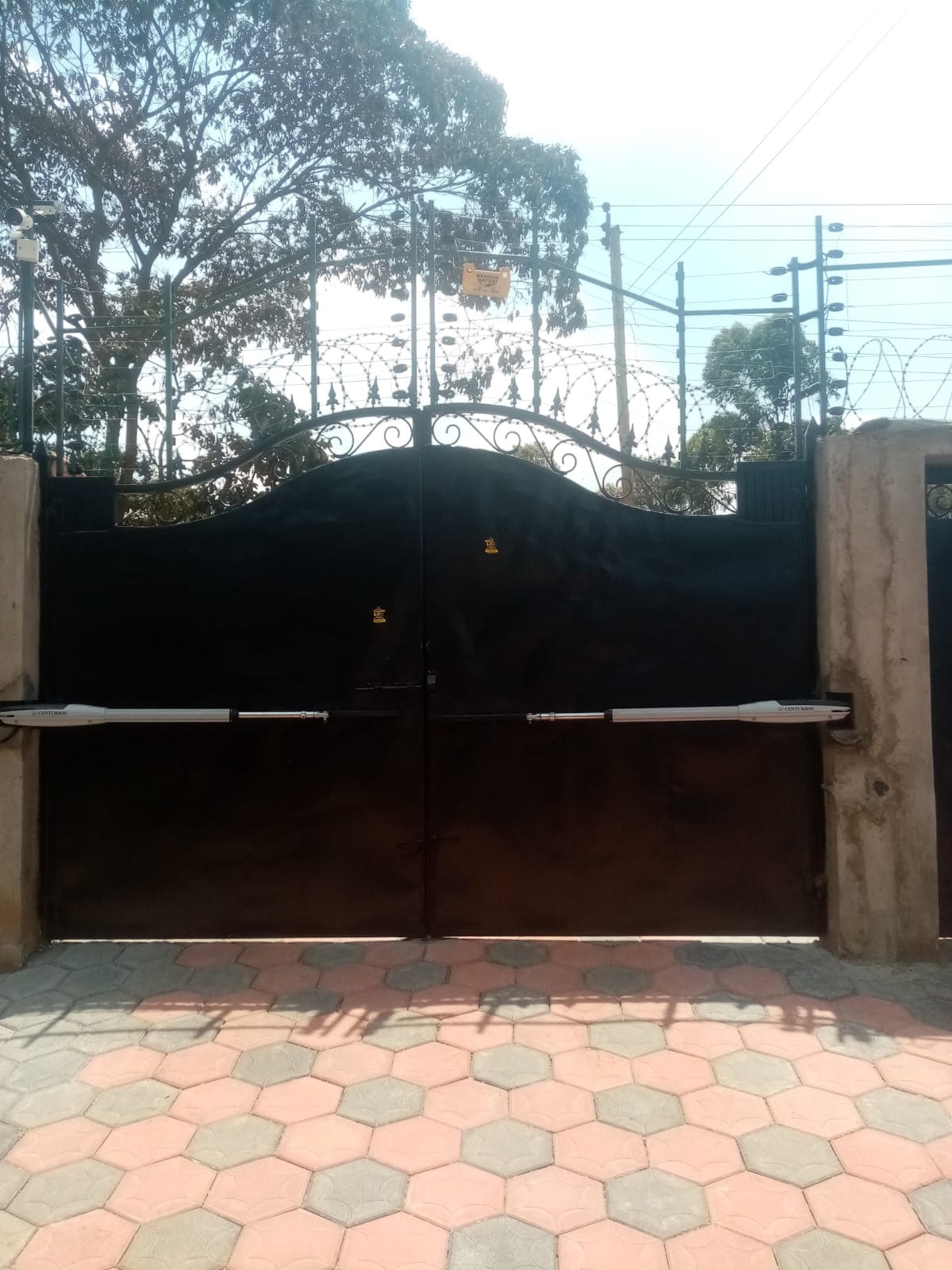 Black double-leaf swing gate with wrought-iron crest, CCTV camera, razor wire, and electric fence at a Karen entrance, wide view