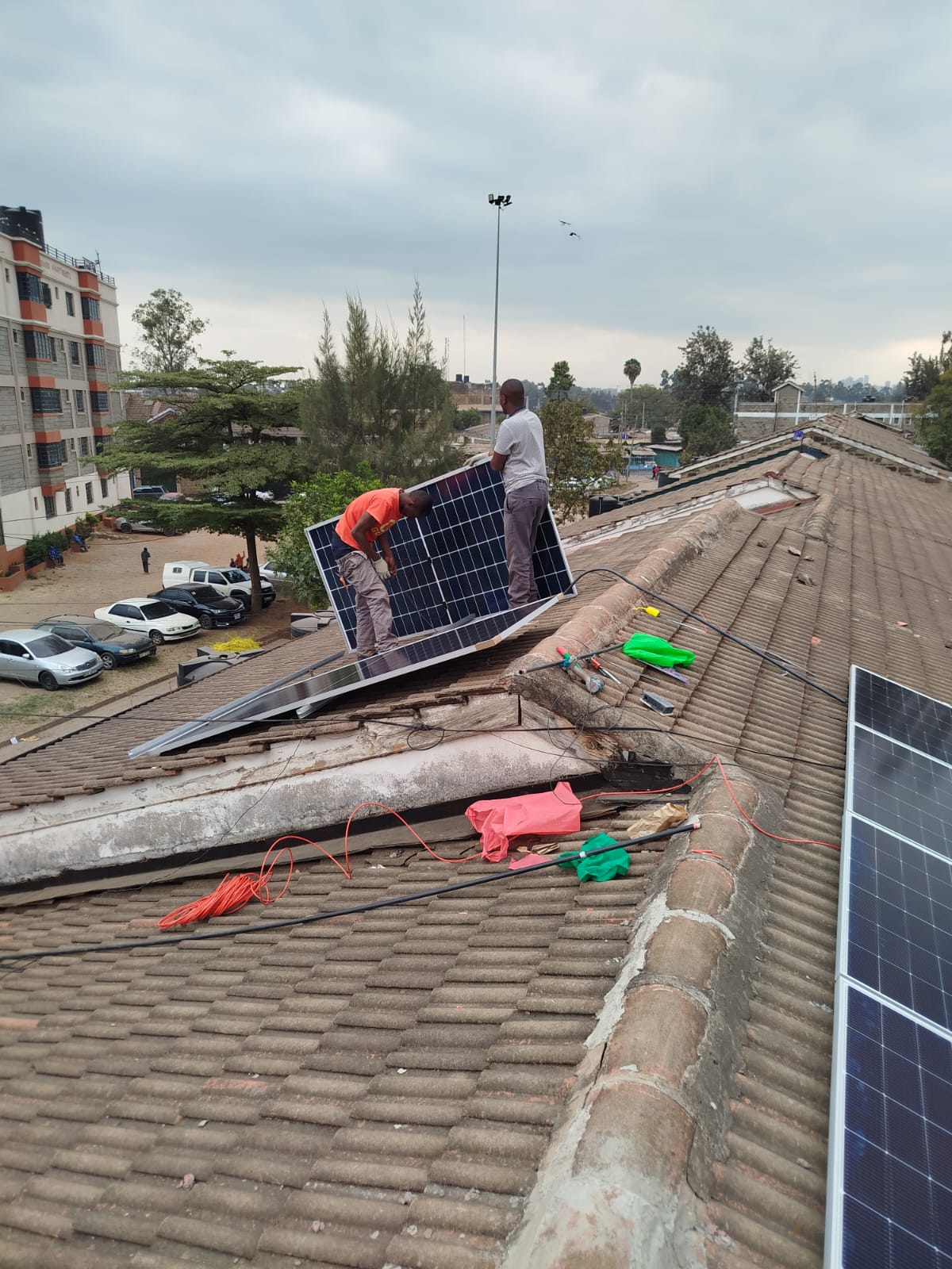 Two technicians lifting a monocrystalline solar panel into position on a tiled roof at a Nairobi apartment complex