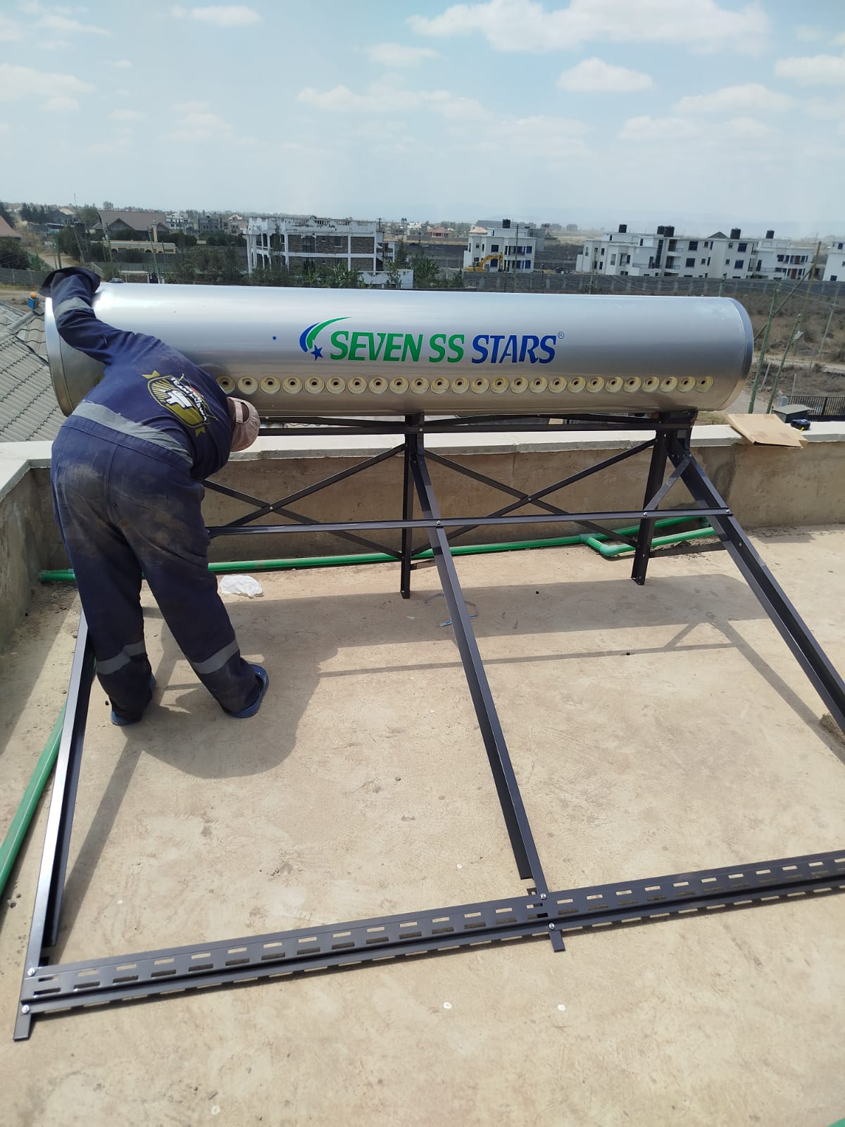 Technician assembling the mounting frame and tank for a 300L solar water heater on a rooftop overlooking a Katani housing development