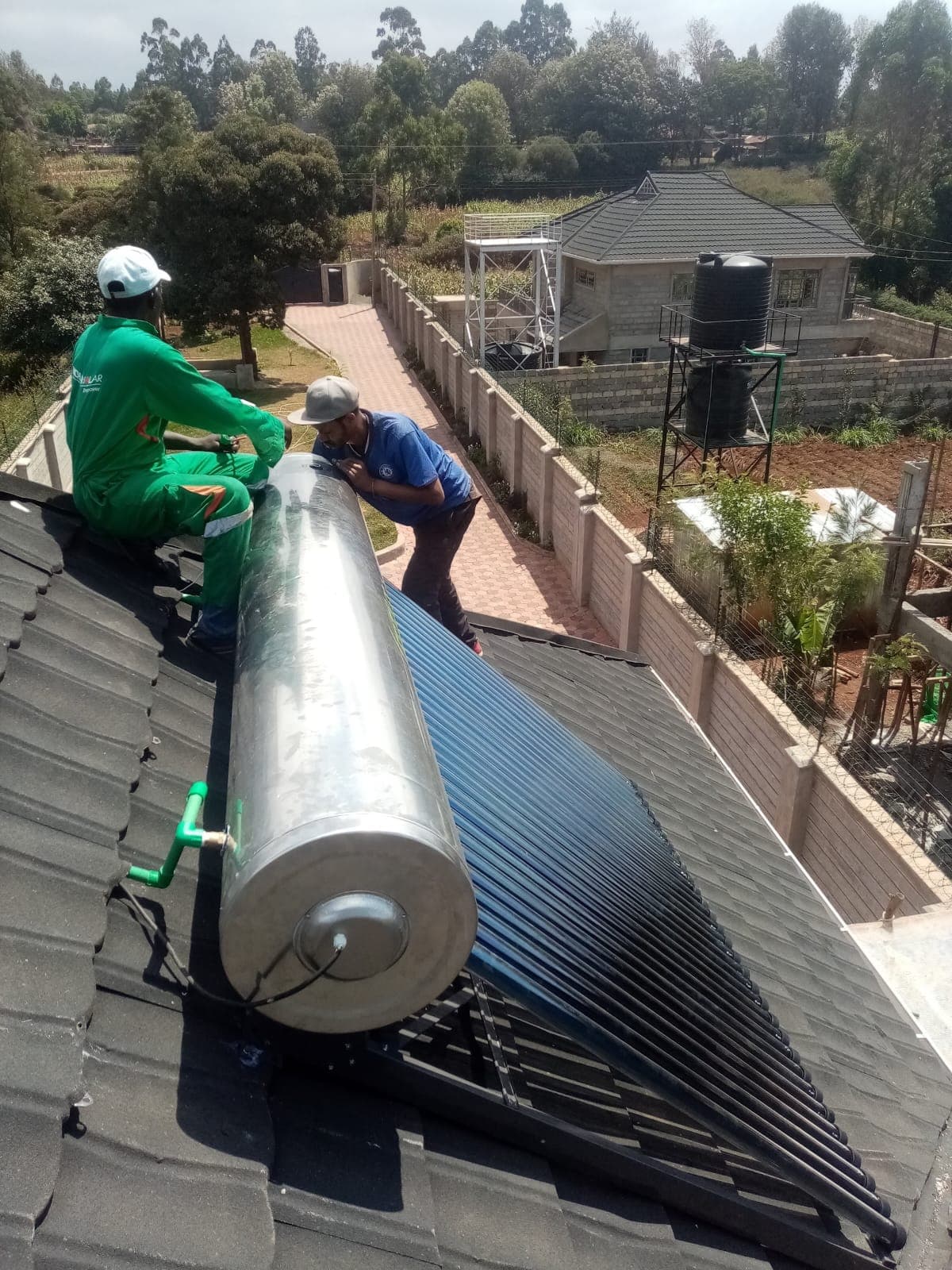 Two technicians installing a 300L evacuated-tube solar water heater tank and collector on a tiled rooftop in Kiambu, with a water tower visible in the neighbouring compound