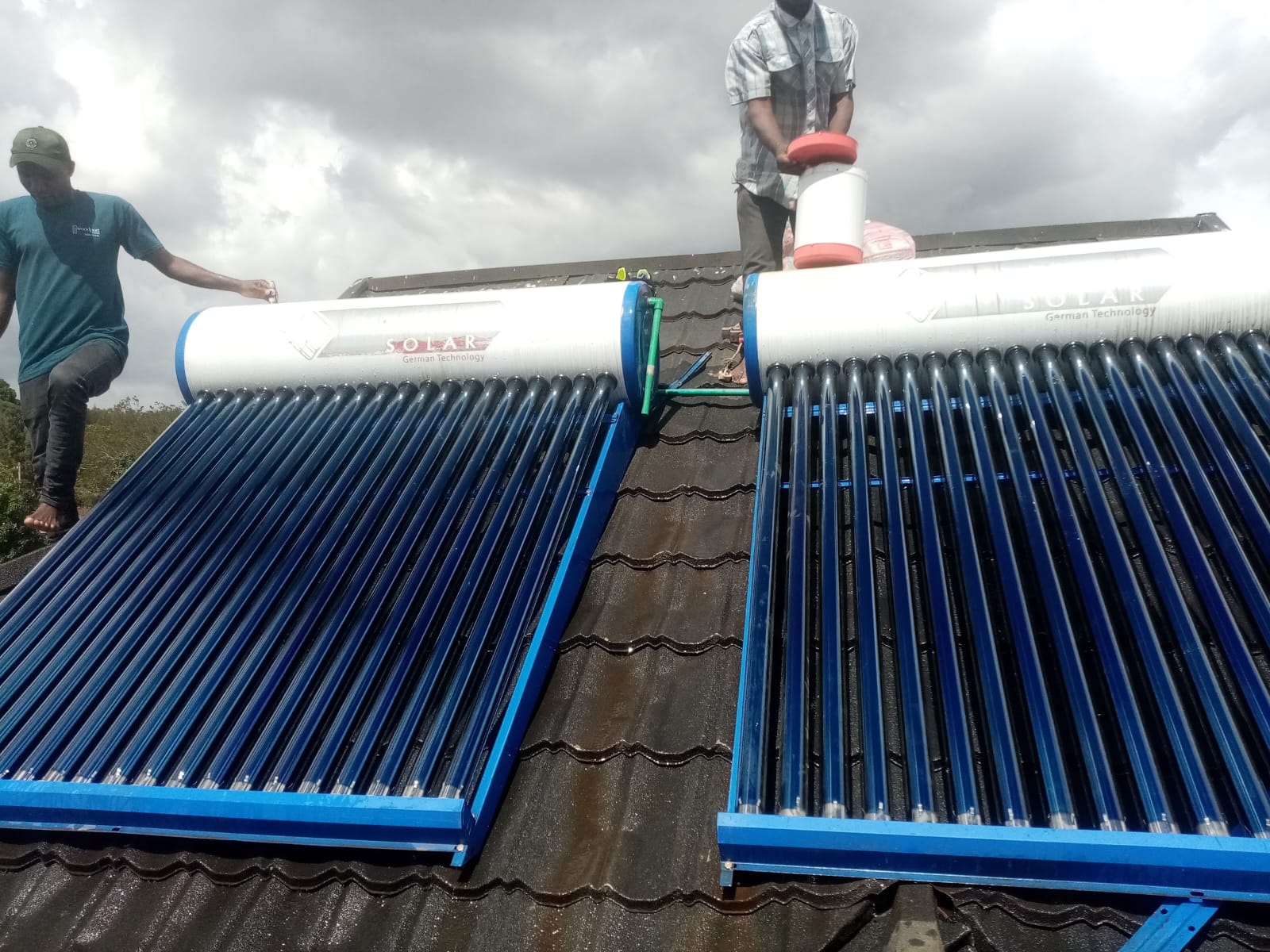 Two technicians working on twin solar water heater tanks installed together on a tiled roof in Kiambu